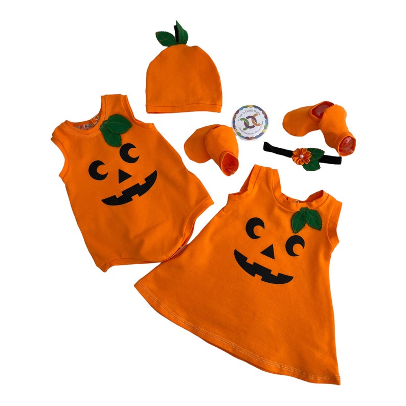 May include: Orange pumpkin-themed baby outfit set with a hat, two pairs of booties, a headband, and two sleeveless shirts. One shirt is a bodysuit and the other is a dress.