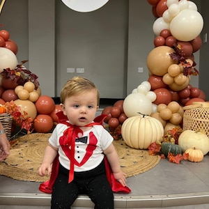 May include: A white short-sleeved bodysuit with a red tuxedo vest graphic, a black bowtie, and three black buttons, paired with a red satin cape tied at the neck and black pants. The outfit is worn by a toddler sitting in front of a balloon arch.