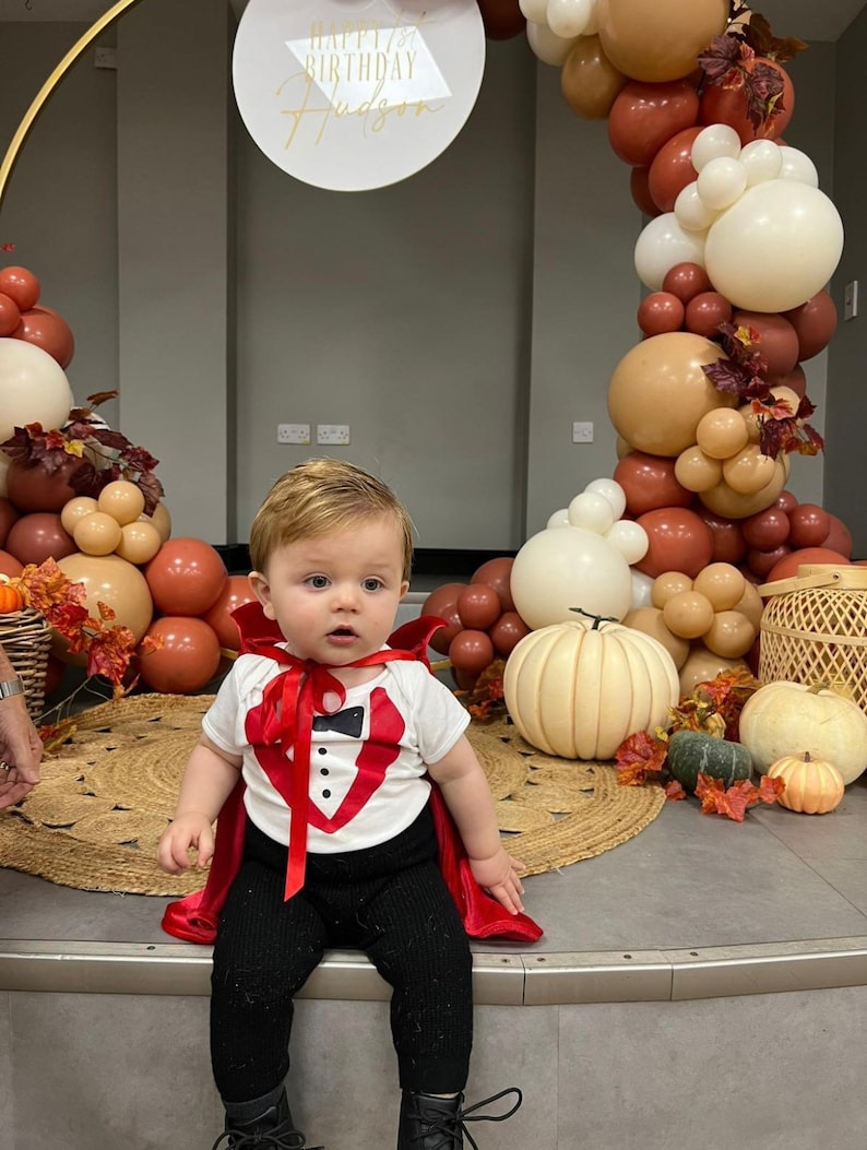 May include: A white short-sleeved bodysuit with a red tuxedo vest graphic, a black bowtie, and three black buttons, paired with a red satin cape tied at the neck and black pants. The outfit is worn by a toddler sitting in front of a balloon arch.