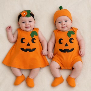 May include: Two infants wearing orange pumpkin-themed outfits. One wears a sleeveless dress with a black jack-o'-lantern face and a matching headband with an orange flower. The other wears a sleeveless romper with a matching face and a pumpkin hat.