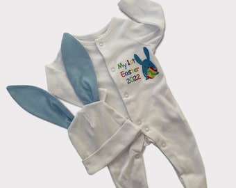 Puede incluir: Mono blanco para bebé con un conejito azul bordado en la parte delantera. El mono dice "My 1st Easter 2022" en letras de colores. Se incluye un gorro de conejito azul a juego.