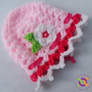 Op de afbeelding: Een roze gehaakte baby muts met een witte bloem en groene bladeren. De muts heeft een roze en witte gehaakte rand.