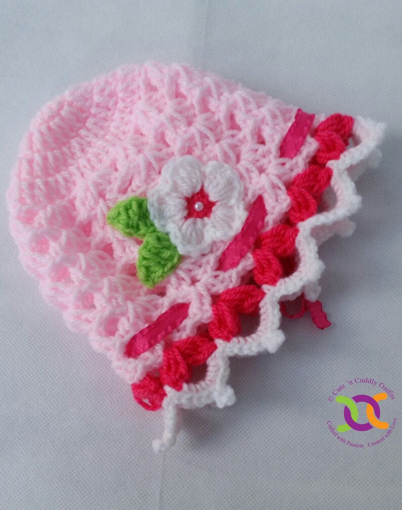 Op de afbeelding: Een roze gehaakte baby muts met een witte bloem en groene bladeren. De muts heeft een roze en witte gehaakte rand.