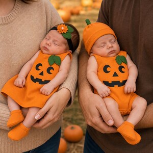 May include: Two infants dressed in orange pumpkin-themed outfits. One wears a sleeveless dress with a black jack-o'-lantern face, green leaf accents, and a matching headband with an orange flower. The other wears a matching onesie and beanie.