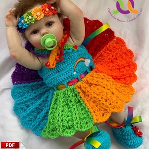 Puede incluir: Un vestido de crochet arco&iacute;ris y zapatos a juego para una mu&ntilde;eca beb&eacute;. El vestido tiene un dise&ntilde;o de arco&iacute;ris con una flor blanca de crochet en la parte delantera. Los zapatos tambi&eacute;n son de crochet y tienen una cinta de arco&iacute;ris.