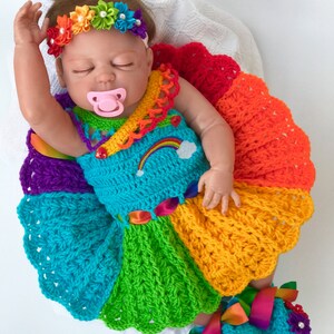 Puede incluir: Una mu&ntilde;eca beb&eacute; con un vestido de crochet arco&iacute;ris con una diadema y zapatos a juego. El vestido tiene un patr&oacute;n de arco&iacute;ris y una nube blanca con un arco&iacute;ris en ella. Los zapatos tambi&eacute;n son de color arco&iacute;ris y tienen una flor en cada uno.