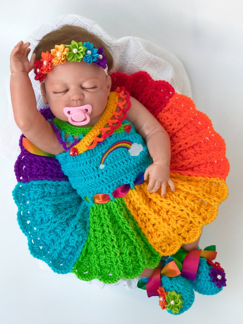 Puede incluir: Una mu&ntilde;eca beb&eacute; con un vestido de crochet arco&iacute;ris con una diadema y zapatos a juego. El vestido tiene un patr&oacute;n de arco&iacute;ris y una nube blanca con un arco&iacute;ris en ella. Los zapatos tambi&eacute;n son de color arco&iacute;ris y tienen una flor en cada uno.