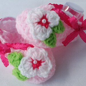 Op de afbeelding: Een paar roze gehaakte babyschoentjes met witte en groene bloemen en roze strikjes. De schoentjes zijn versierd met kleine witte pareltjes.