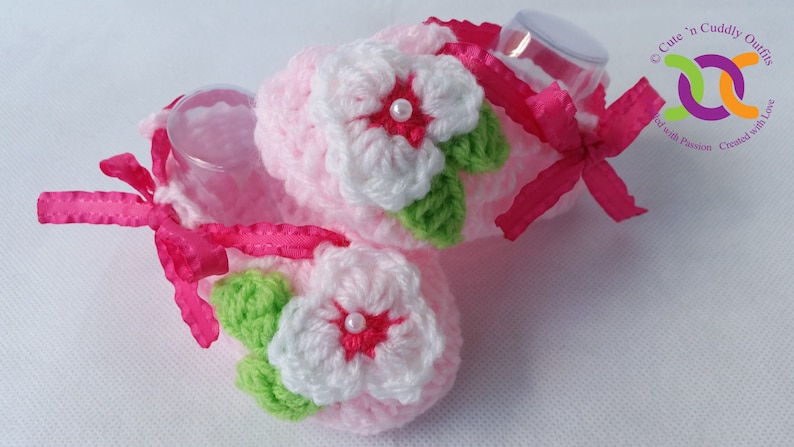 Op de afbeelding: Een paar roze gehaakte babyschoentjes met witte en groene bloemen en roze strikjes. De schoentjes zijn versierd met kleine witte pareltjes.