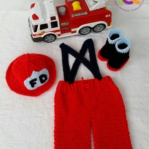 May include: A red and white toy fire truck with a ladder is next to a crocheted firefighter outfit. The outfit includes red pants with suspenders, a matching hat with "FD" on it, and black and blue booties.
