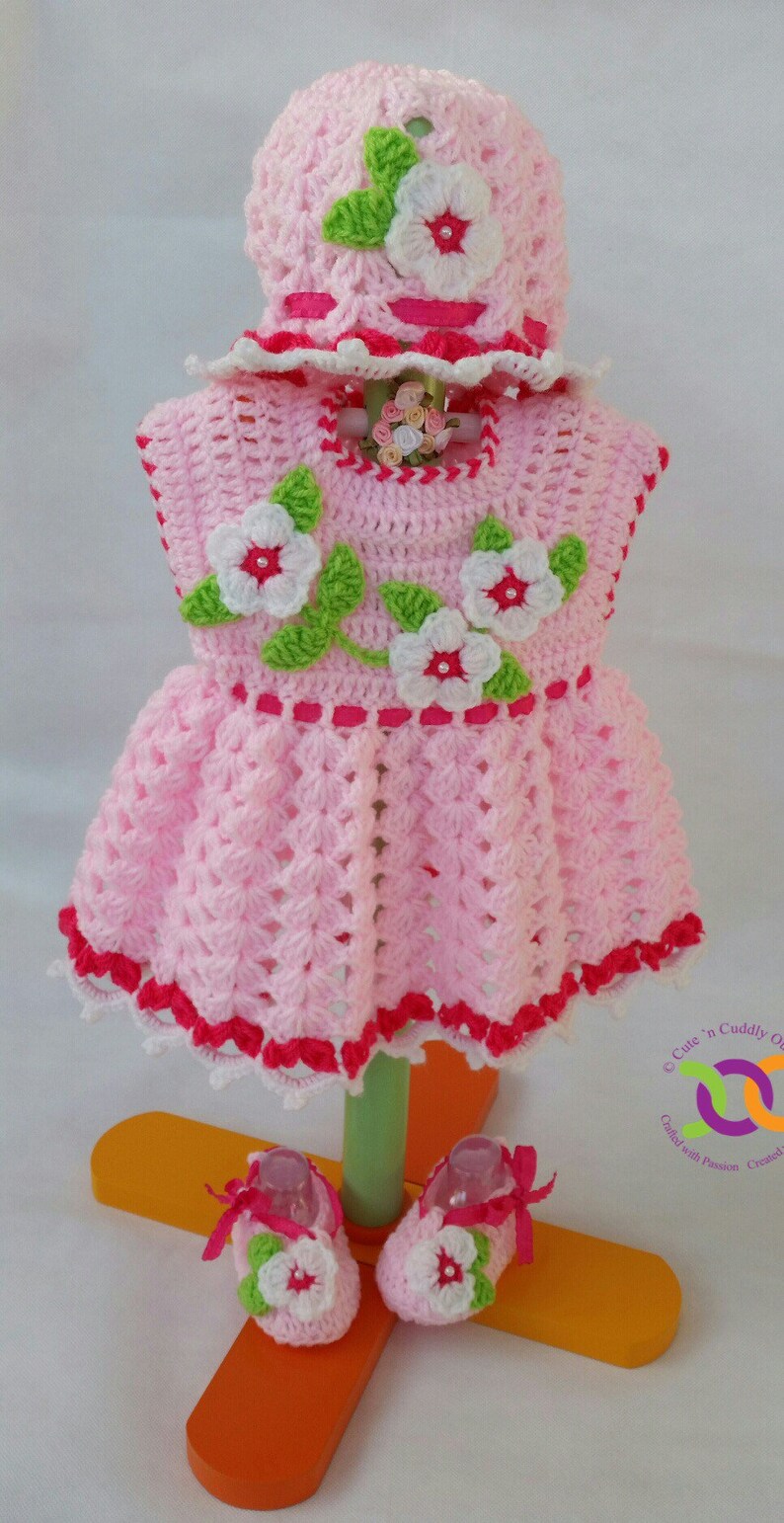 Op de afbeelding: Een roze gehaakt babyjurkje met witte en groene bloemenaccenten, een bijpassende muts en roze en witte gehaakte schoentjes. Het jurkje en de schoentjes hebben een rode bies.