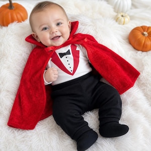 May include: A baby outfit consisting of a white long-sleeve shirt with a printed red tuxedo vest, black bowtie, and buttons, paired with solid black footed pants and a bright red velvet cape that features a high collar and a red ribbon tie.