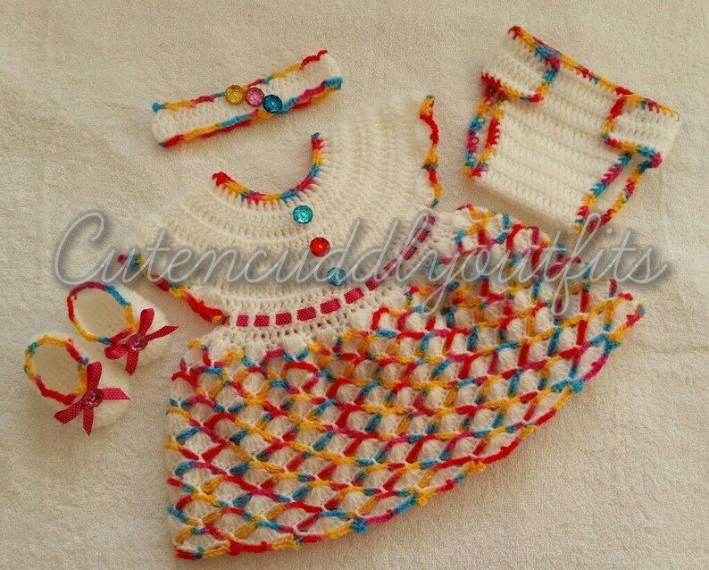 May include: A crocheted baby outfit set in white with colorful accents. The set includes a dress, a headband, booties, and a diaper cover. The dress has a colorful geometric pattern and is decorated with colorful buttons. The headband is decorated with colorful buttons. The booties are white with red bows. The diaper cover is white with colorful trim.