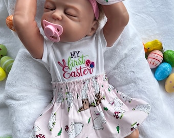Könnte beinhalten: Eine Baby-Puppe trägt einen weißen Strampler mit dem Text "My first Easter" und einen rosa Rock mit Hasenmuster. Die Puppe trägt ein rosa Stirnband mit Hasenohren. Die Puppe ist von bunten Ostereiern umgeben.