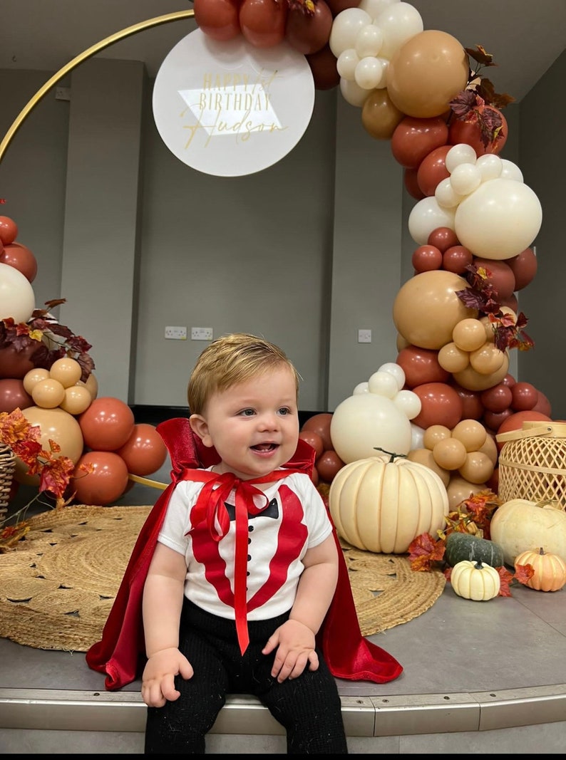 May include: A child wearing a white shirt with a red tuxedo print, a red cape, and black pants. Behind the child is a gold circular frame decorated with brown, cream, and white balloons, artificial autumn leaves, and a sign reading Happy 1st Birthday Hudson.
