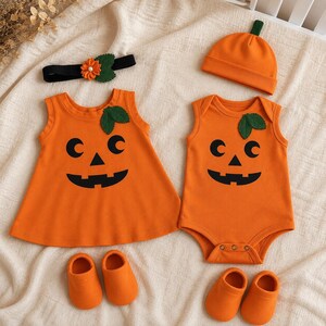 May include: An orange sleeveless dress and an orange sleeveless bodysuit, both featuring a black jack-o'-lantern face and green leaf accents. The set includes an orange beanie with a green stem, a black headband with a flower, and two pairs of orange booties.