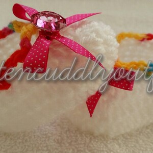 May include: White crocheted baby shoes with a pink bow and a sparkly pink gem. The shoes have a colorful crocheted trim.