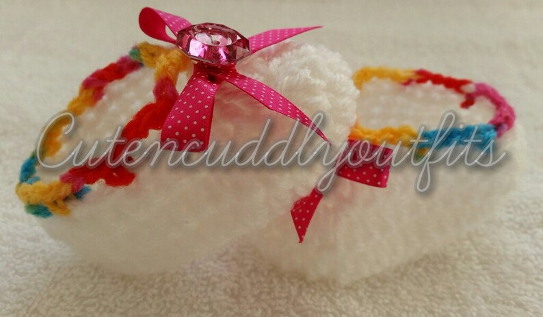 May include: White crocheted baby shoes with a pink bow and a sparkly pink gem. The shoes have a colorful crocheted trim.