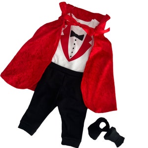 May include: A white bodysuit featuring a red tuxedo graphic with a black bowtie and buttons, paired with black pants, a red velvet cape with a ribbon tie, and two black fabric booties. The ensemble creates a formal look with a vibrant red cape.