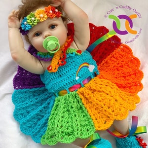 Puede incluir: Una mu&ntilde;eca beb&eacute; con un vestido de crochet arco&iacute;ris con una diadema y botines a juego. El vestido tiene una cinta arco&iacute;ris atada alrededor de la cintura y los botines tienen cintas arco&iacute;ris. La mu&ntilde;eca lleva un chupete.