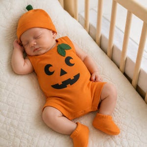 May include: A bright orange sleeveless baby bodysuit featuring a black jack-o'-lantern face print and two green fabric leaves on the shoulder. The outfit includes a matching orange beanie with a small green stem and a pair of solid orange booties.
