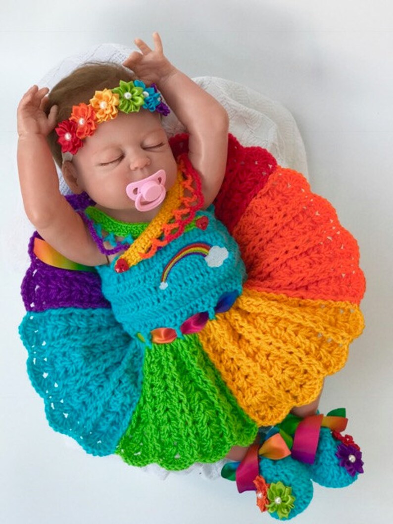 Puede incluir: Una mu&ntilde;eca beb&eacute; con un vestido de crochet arco&iacute;ris con una diadema y zapatos a juego. El vestido est&aacute; hecho de hilo de diferentes colores, incluyendo rojo, naranja, amarillo, verde, azul, &iacute;ndigo y violeta. La mu&ntilde;eca lleva un chupete rosa.