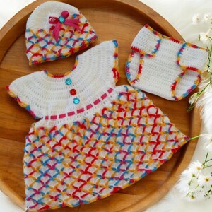 May include: A crocheted baby outfit set in white with colorful accents. The set includes a dress, a hat, and a diaper cover. The dress has a colorful geometric pattern and a pink ribbon around the neck. The hat has a pink ribbon with a jewel accent. The diaper cover has a colorful trim.