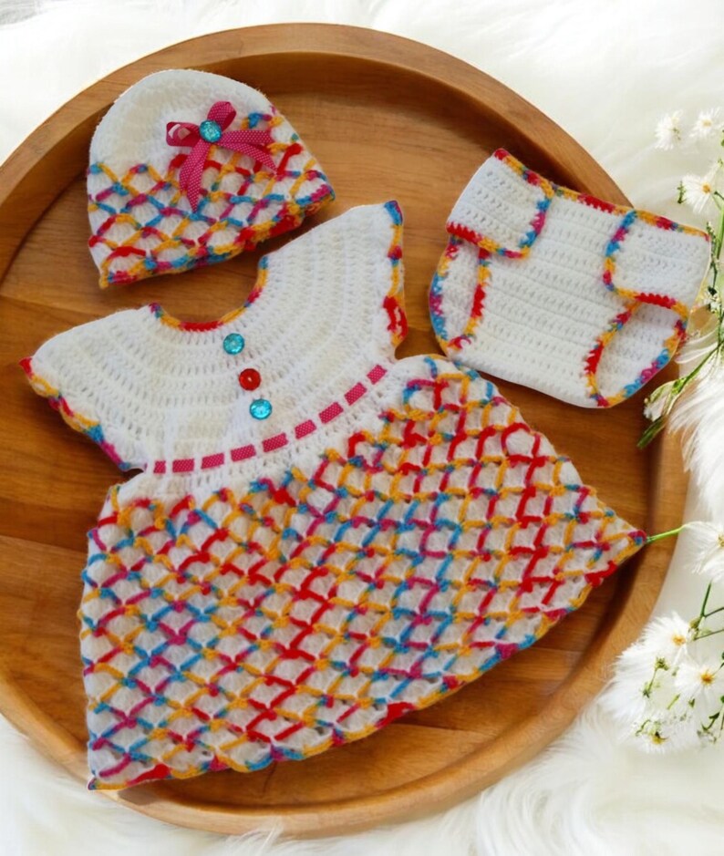 May include: A crocheted baby outfit set in white with colorful accents. The set includes a dress, a hat, and a diaper cover. The dress has a colorful geometric pattern and a pink ribbon around the neck. The hat has a pink ribbon with a jewel accent. The diaper cover has a colorful trim.
