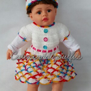 May include: A white crocheted dress with a colorful geometric pattern, a pink ribbon, and colorful buttons. The dress is worn by a doll with brown curly hair and a white headband with colorful stripes. The doll is wearing white crocheted shoes with pink bows and rhinestones.
