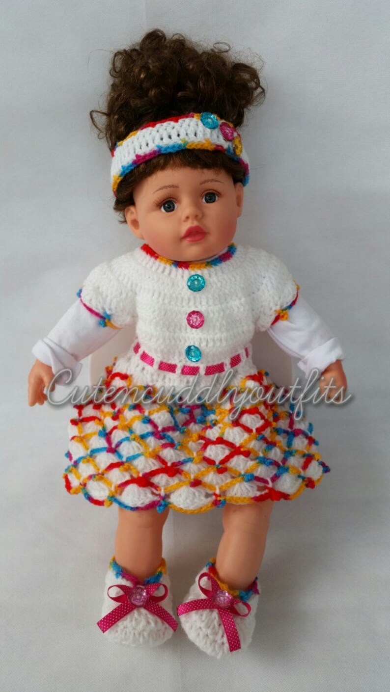 May include: A white crocheted dress with a colorful geometric pattern, a pink ribbon, and colorful buttons. The dress is worn by a doll with brown curly hair and a white headband with colorful stripes. The doll is wearing white crocheted shoes with pink bows and rhinestones.
