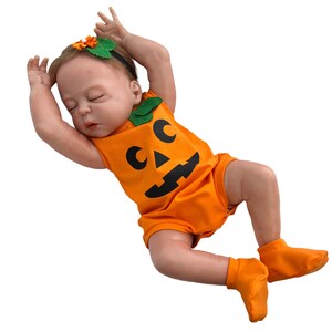 May include: A baby doll wearing an orange pumpkin costume with a black jack-o'-lantern face. The doll is wearing a green leaf headband and orange booties.