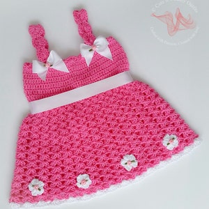 May include: A pink crocheted baby dress with white straps, a white ribbon around the waist, and white crocheted flowers around the hem.