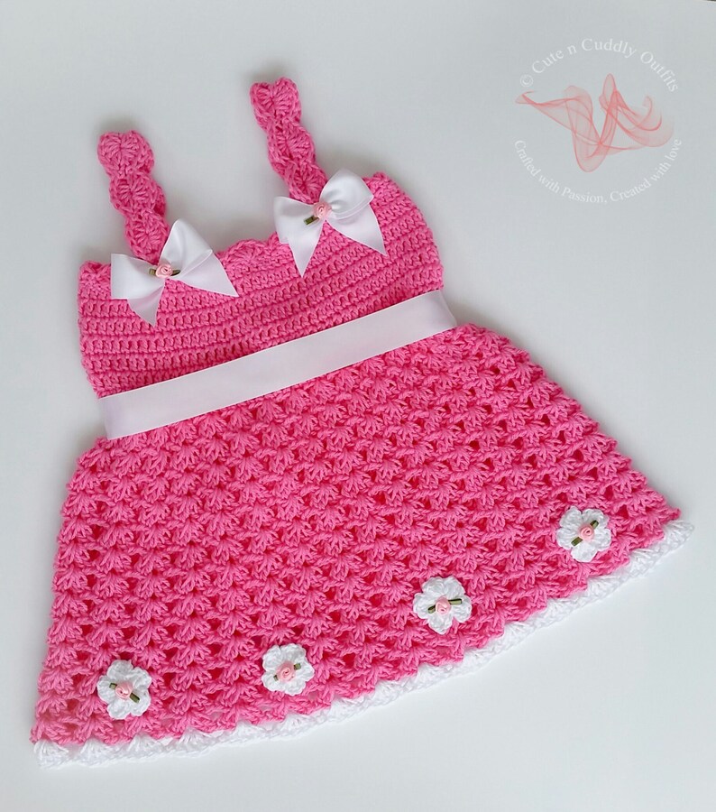 May include: A pink crocheted baby dress with white straps, a white ribbon around the waist, and white crocheted flowers around the hem.