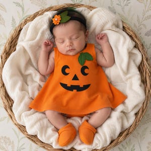 May include: A baby wearing an orange sleeveless dress with a black jack-o'-lantern face print and a green leaf accent. The baby also wears a black headband with an orange flower and green leaves, along with matching orange booties.