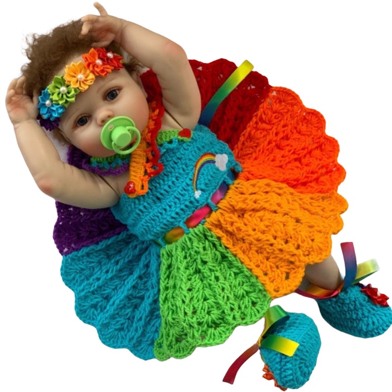 Puede incluir: Una mu&ntilde;eca beb&eacute; con un vestido de crochet arco&iacute;ris con zapatos a juego. El vestido tiene un dise&ntilde;o de arco&iacute;ris y una nube blanca con un arco&iacute;ris en ella. La mu&ntilde;eca lleva una diadema con flores de colores.