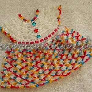 May include: A white crocheted baby dress with a colorful geometric pattern and a pink, red, and blue trim. The dress has a round neckline and short sleeves. There are three decorative buttons on the front of the dress.