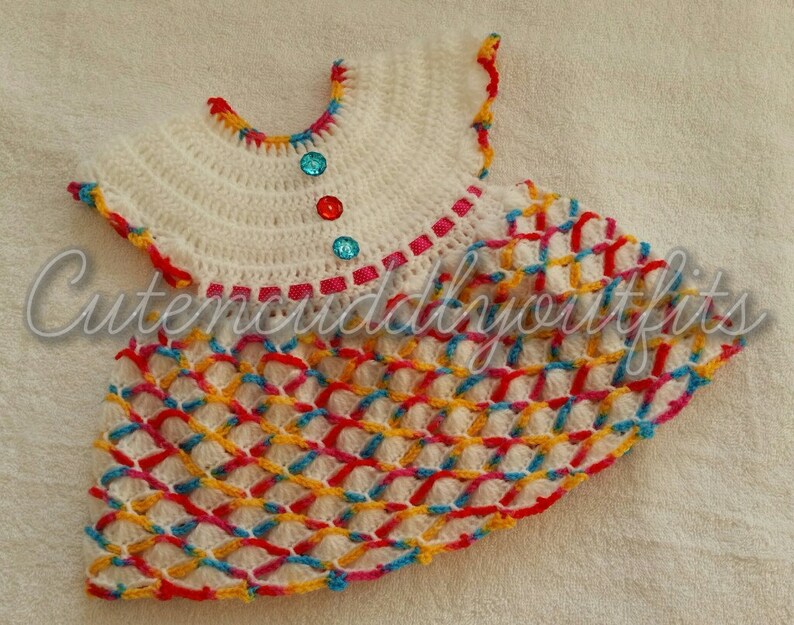 May include: A white crocheted baby dress with a colorful geometric pattern and a pink, red, and blue trim. The dress has a round neckline and short sleeves. There are three decorative buttons on the front of the dress.