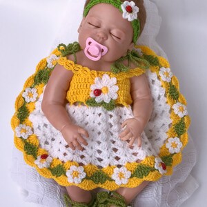 Może przedstawiać: Sukienka dla lalki wykonana na szydełku, z białym tłem, ż&oacute;łtymi i zielonymi akcentami oraz detalami w postaci stokrotek i biedronek. Sukienkę nosi lalka z zieloną opaską na głowę i r&oacute;żowym smoczkiem.