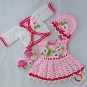 Op de afbeelding: Een roze gehaakte babyjurk met witte en groene bloemenaccenten, een wit gehaakt vest met roze bies, een roze gehaakte muts met witte bies en een paar witte gehaakte slofjes met roze bies.