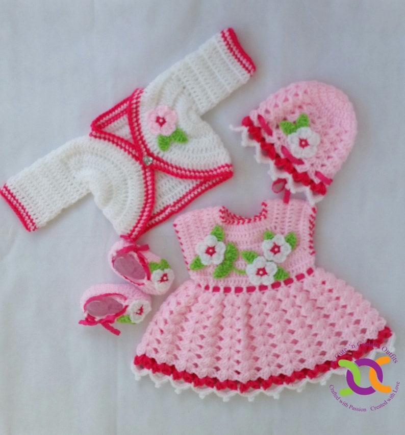 Op de afbeelding: Een roze gehaakte babyjurk met witte en groene bloemenaccenten, een wit gehaakt vest met roze bies, een roze gehaakte muts met witte bies en een paar witte gehaakte slofjes met roze bies.