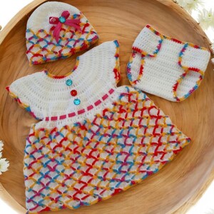 May include: A white crocheted baby outfit with a rainbow pattern. The outfit includes a dress, a hat, and a diaper cover. The dress has a white bodice with pink and red trim and a rainbow crocheted skirt. The hat is white with a pink bow and rainbow crocheted trim. The diaper cover is white with rainbow crocheted trim.
