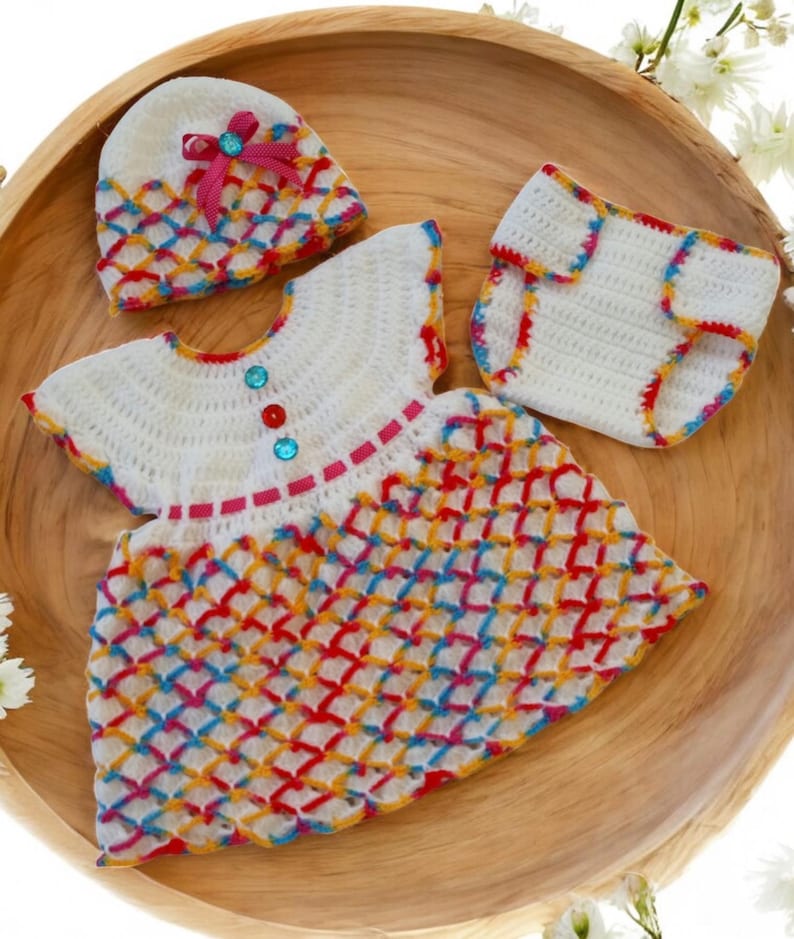 May include: A white crocheted baby outfit with a rainbow pattern. The outfit includes a dress, a hat, and a diaper cover. The dress has a white bodice with pink and red trim and a rainbow crocheted skirt. The hat is white with a pink bow and rainbow crocheted trim. The diaper cover is white with rainbow crocheted trim.