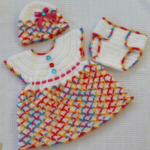 May include: A white crocheted baby outfit with a matching hat. The dress and diaper cover have a colorful diamond pattern with red, yellow, blue, and pink accents. The hat has a pink bow.