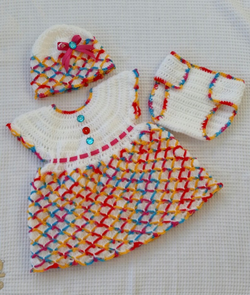 May include: A white crocheted baby outfit with a matching hat. The dress and diaper cover have a colorful diamond pattern with red, yellow, blue, and pink accents. The hat has a pink bow.