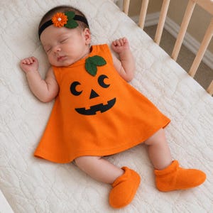 May include: An infant wearing an orange sleeveless dress with a black jack-o'-lantern face print and two green felt leaves at the neckline. The infant also wears matching orange booties and a black headband featuring an orange flower and green leaves.