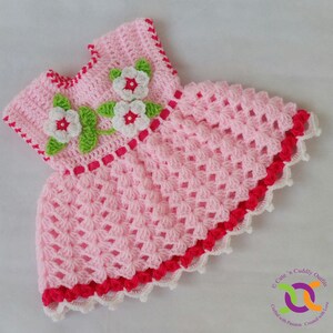 Op de afbeelding: Een roze gehaakt babyjurkje met witte en rode bies. Het jurkje heeft een bloemenmotief op het bovenlijf met witte bloemen en groene bladeren.