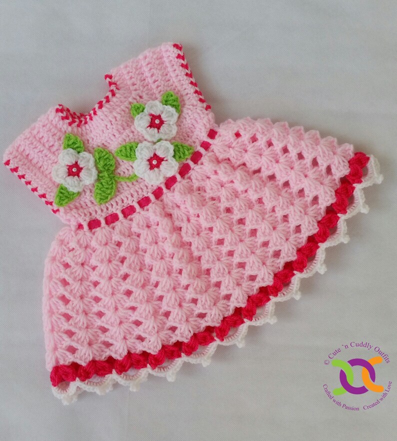 Op de afbeelding: Een roze gehaakt babyjurkje met witte en rode bies. Het jurkje heeft een bloemenmotief op het bovenlijf met witte bloemen en groene bladeren.