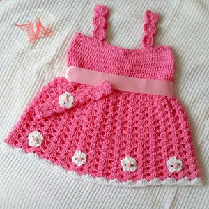 May include: A pink crocheted baby dress with white crocheted flowers and a pink satin ribbon. The dress has a scalloped neckline and a white crocheted trim.