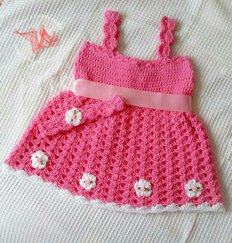 May include: A pink crocheted baby dress with white crocheted flowers and a pink satin ribbon. The dress has a scalloped neckline and a white crocheted trim.