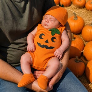 May include: An orange baby bodysuit featuring a black jack-o'-lantern face print and a green fabric leaf accent at the neckline. The outfit includes a matching orange beanie cap and soft orange booties, creating a complete pumpkin-themed ensemble.